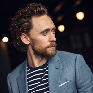 Tom Hiddleston, Caucasian male actor, in a serious pose with a full beard and curly medium-brown hair. Wears a stylish striped shirt and matching blazer. Set against a dark background with soft lighting creating a gentle glow. Close-up, high-resolution photo with shallow depth of field, emphasizing his features.