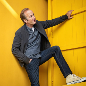 Bob Odenkirk, with balding hairline, dressed in casual attire, posing dynamically against a bright yellow backdrop.