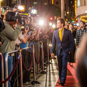 Matt Bomer at a bustling Hollywood premiere. Wearing a tailored navy suit and yellow shirt. Surrounded by flashing cameras and a crowd of excited fans.