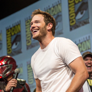 Chris Pratt, smiling brightly, wearing a casual white t-shirt. On stage at a Comic-Con event, with fans and cosplayers in the background.