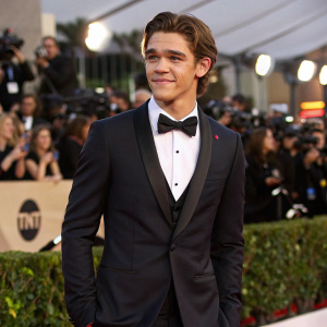 Brenton Thwaites, actor, in elegant black suit and bow tie, at the glamorous evening awards ceremony. The background is a sea of flashing cameras and excited crowd.