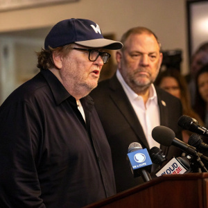 Photograph of Michael Moore and Harvey Weinstein at a controversial press conference. Michael Moore in dark shirt, standing quietly in background. Harvey Weinstein, bearded and wearing a white shirt with a dark suit, addresses the crowd from the podium with intensity.