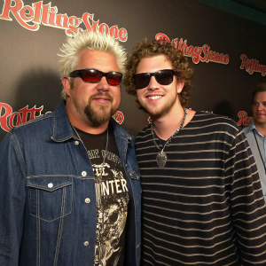 Guy Fieri and Hunter Fieri posing for a candid shot. Guy in denim jacket, graphic tee, and shades. Hunter in striped shirt. Rolling Stone logo backdrop