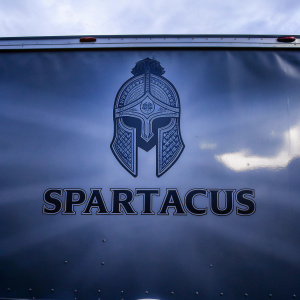 Spartacus logo featuring a detailed warrior helmet design in blue color. The word Spartacus is displayed below in bold letters. A rugged trailer with the same Spartan theme against a dark backdrop.