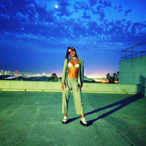 Karrueche Tran, radiant with a vibrant smile, adorned in eclectic jewelry and body art, standing confidently against a sleek urban backdrop.