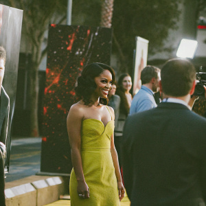 Cynthia Addai-Robinson on the red carpet at the 'Spartacus: War of the Damned' premiere. Wearing a bright yellow gown, she smiles for the cameras amidst the bustling event atmosphere. The backdrop features stark black with bold red splatters and event branding.