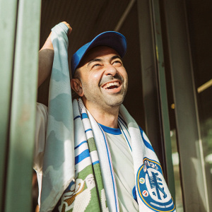 Roberto Di Matteo, with short hair, wearing Chelsea FC jersey and cap. He looks ecstatic. Dark background