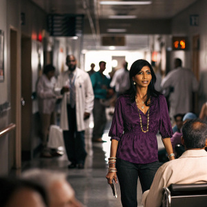 Reshma Shetty as Dr. Divya Katdare from 'Royal Pains'. Wearing a vibrant purple blouse and gold jewelry. Backdrop is a bustling hospital hallway.