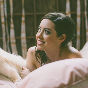 Aubrey Plaza with a warm smile, minimal makeup, and stylish dark hair. Close-up shot against a pastel pink backdrop.