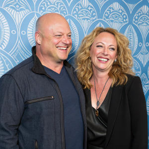 Michael Chiklis and Virginia Madsen attend the SAG-AFTRA Foundation Benefit Gala. Smiling widely, casual attire, in front of blue patterned backdrop.