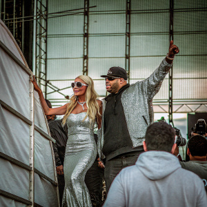 Fat Joe and Paris Hilton at a bustling music festival. Paris, blonde, wears a dazzling silver gown. Fat Joe in a black cap and stylish sunglasses.