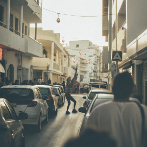 Antiparos Island, Greece. A bustling modern street with cars and people. Brightly colored buildings and neon signs. The cat is replaced by a street performer.