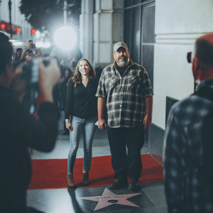 Cara Whitney and Larry the Cable Guy at the Hollywood Walk of Fame. Black shirt and jeans, plaid shirt and cap. Crowds and camera flashes in background.
