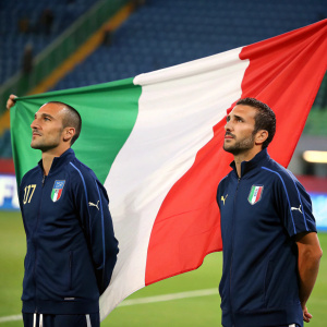 Fabio Cannavaro and Gianluca Zambrotta standing beside a giant Italian flag. Solemn expressions. Stadium background.