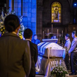 A solemn photo of a funeral ceremony at Manila Cathedral. Kris Aquino is present, focusing on the casket draped in white. Attendees are in formal attire, with subdued lighting highlighting the ceremony's somber tone.
