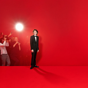 Timothée Chalamet in a candid shot from a film premiere. Wearing a black suit and bow tie. The vibrant red carpet backdrop is bustling with photographers and fans.