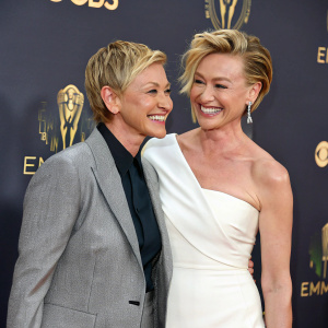 Ellen DeGeneres and Portia De Rossi at a gala event. Both smiling broadly. Portia in a white gown, Ellen in a grey blazer. Dark backdrop with illuminated logos
