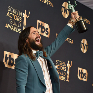 Jared Leto celebrating at the Screen Actors Guild Awards. Holding a golden statuette, excited smile. Dark backdrop with event logos and sculptural elements.