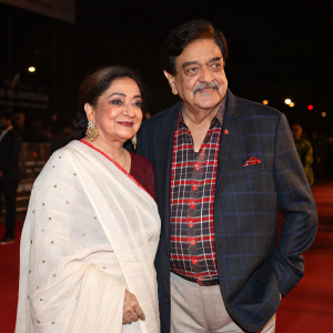 Stalwart Bollywood actor Shatrughan Sinha and Indian cinema icon Poonam Sinha at the red carpet event. Shatrughan, in a tartan shirt and blazer, Poonam in a white saree with a traditional red bindi. Dark backdrop.
