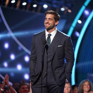 Aaron Rodgers, dressed in a formal suit, at the ESPY Awards ceremony. Stage background with bright lights and cheering crowd