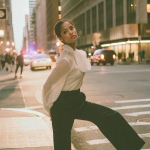 Gugu Mbatha-Raw in a chic white blouse and black pants, posing on a busy city street at sunset.