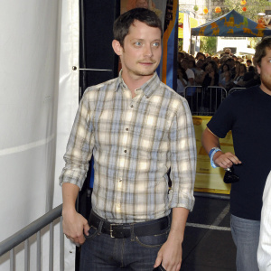 Elijah Wood, dressed casually in a plaid shirt, poses for a candid shot at a Hollywood street festival. Blurred crowd in the background