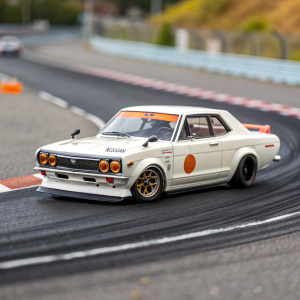 A detailed photograph of a 1/32 scale white Nissan Skyline KPGC10 model car with orange highlights racing on a track. The model showcases the exquisite craftsmanship and features of the club series kit, alongside the proud Nissan logo.