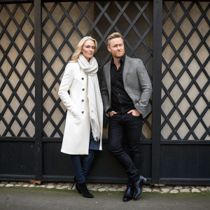 Storm and Ronan Keating posing for a casual photo shoot. Lattice-patterned backdrop in dark tones. Storm in white coat and scarf, Ronan in black shirt, grey blazer, dark pants.