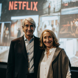 Sam Waterston and Lynn Louisa Woodruff attending a press conference for their new Netflix series. They are standing in front of a large, colorful backdrop featuring the Netflix logo and show graphics. Sam is dressed in a dark suit while Lynn is in a stylish, layered blouse. They both look directly at the camera with smiles.