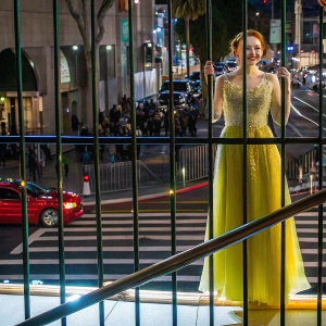Madelaine Petsch, the fair-skinned actress, wearing a vibrant yellow dress with intricate lace detailing, posing in front of a bustling street scene.