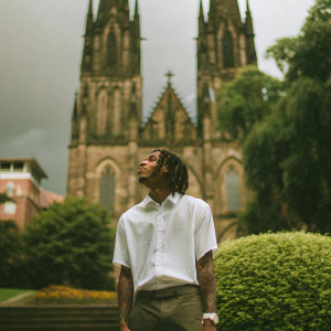Playboi Carti, rapper with distinctive dreads, standing in front of a gothic cathedral. Dark sky, stormy weather