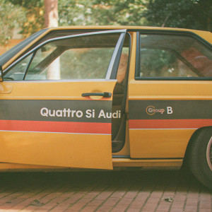 Vintage Audi Quattro S1 Areo rally car. Classic yellow and black color scheme. German flag design on doors. 'Quattro Si Audi' and 'Group B' text on body. Modern design elements
