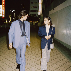 Anthony Eden and Clarissa Eden, dressed in formal attire, walking on a busy street at night. Bright neon signs and modern buildings in the background.