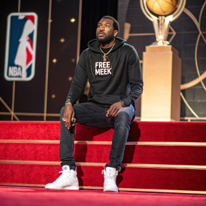 Meek Mill, male artist of African descent, intense expression, wearing a black hoodie with 'Free Meek' slogan, dark jeans, and white sneakers. NBA Awards stage background.