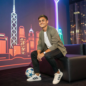 Paulo Dybala, male soccer player, posing in a casual outfit at a fan meet. The backdrop is a city skyline with neon lights.