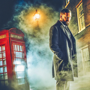 Tom Hardy as James Delaney, from the show Peaky Blinders. Wears a dark suit with a beard, standing in front of a foggy London street backdrop.