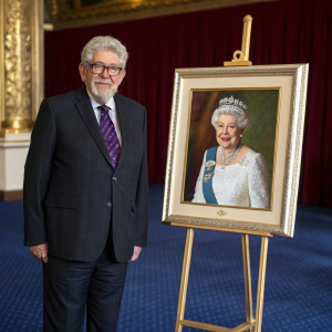 Rolf Harris, with his iconic beard and glasses, standing beside a portrait of the Queen. Formal setting with royal blue carpet.