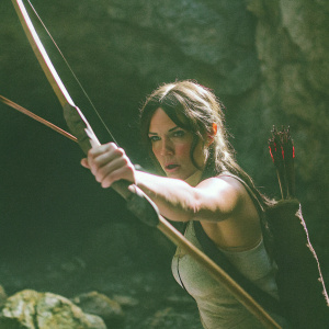A fierce archer, resembling Lara Croft, stands poised with bow drawn. Her brown hair cascades behind her. Dark, mysterious cave background.