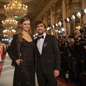 Fernando Alonso and Dasha Kapustina at a glamorous fashion gala, surrounded by luxurious decor and flashing cameras.
