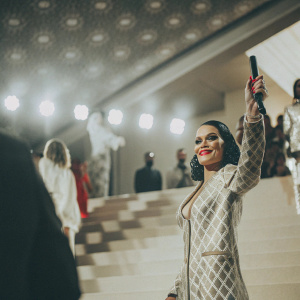 Jessie J at the 2022 Met Gala, vibrant red lipstick, patterned dress, posing confidently on a stage with streaming lights and glamorous crowd.