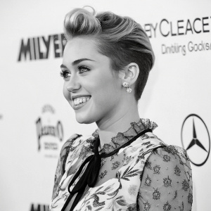 Miley Cyrus, with an upbeat expression, captured in a close-up monochrome photograph. She wears a floral print blouse with intricate lace detailing and a stylish updo. The backdrop features vibrant logos and branding against a solid white background.