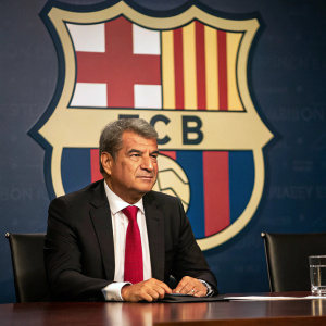 Joan Laporta, FC Barcelona president, in a stern pose. Wearing a tailored black suit and red tie. Sitting at a conference table with the club's logo prominently displayed behind him.