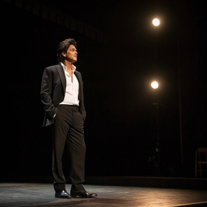 Shah Rukh Khan, with intense expression, wearing a black suit and white shirt. Empty stage, single spotlight, dimly lit