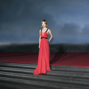 Caucasian actress Emily Browning in elegant evening gown. Short, layered bob, fair skin, subtle makeup. Vibrant red gown with ruched bodice. Dark, blurred background with reddish hues. High fashion magazine photo.