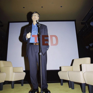 Bill Gates speaking at TED Talks. Dark suit, blue shirt. White backdrop with TED logo. Stage lights illuminate his focused expression.