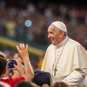 Francis, the Pope, smiling warmly. Crowded stadium, people waving back. Background blurred