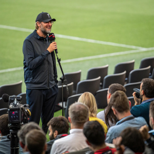 Jurgen Klopp, in a casual outfit, speaking at a press conference. Background: audience and reporters.