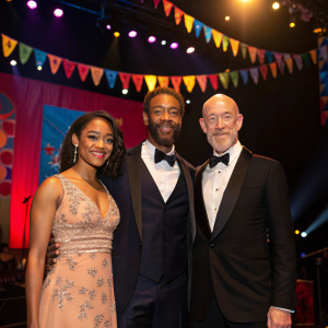 Yasmine Delawari Johnson, Matthew Johnson, and J.K. Simmons at a charity gala. Brightly lit stage with banners.