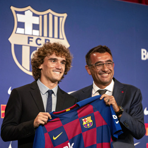 Antoine Griezmann and Josep Maria Bartomeu at a press conference. Griezmann with curly hair, holding a Barcelona FC jersey. Bartomeu in a formal suit, standing behind him. Background features the club's logo and banners.