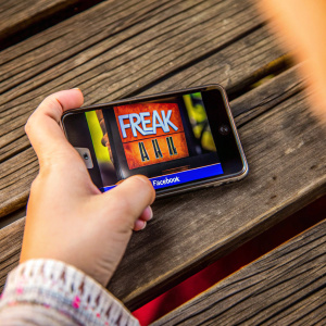 A close-up photo of a hand holding a smartphone showcasing the game 'Freak Jack' with Facebook logo. The smartphone is placed on a rustic wooden table.